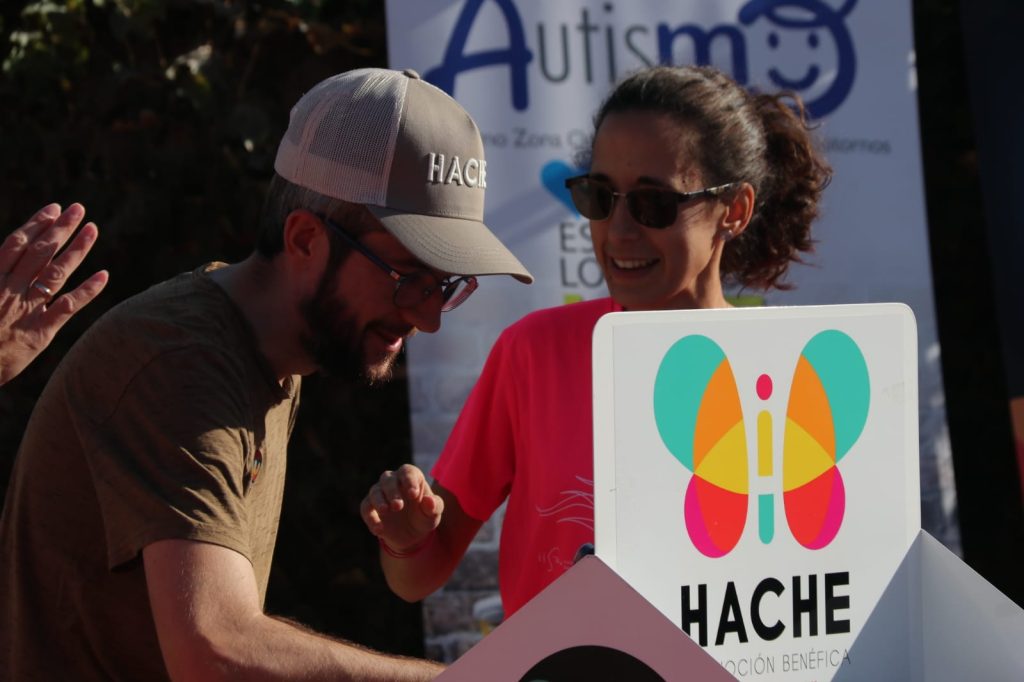 Dos personas del equipo hablando en el fondo. En primer plano, se ve la pegatina del logo de Hache en grande, colocada en la estantería del stand