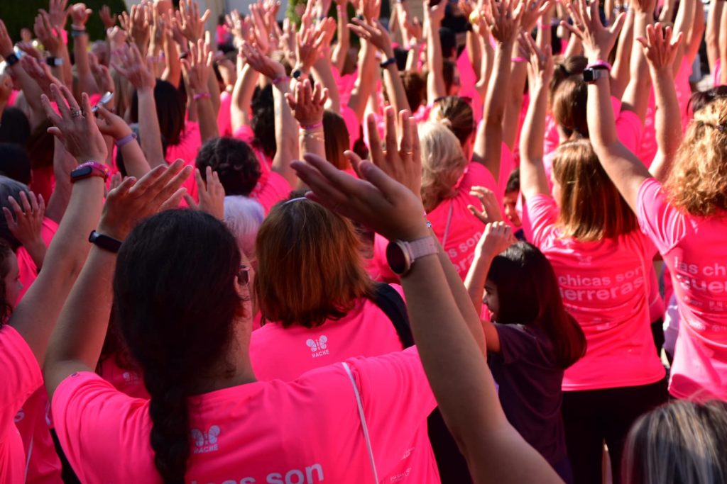 Muchas mujeres de la carrera de espaldas con las manos hacia arriba. Se ve en primer plano la trasera de la camiseta de una de ellas, con el logo de Hache