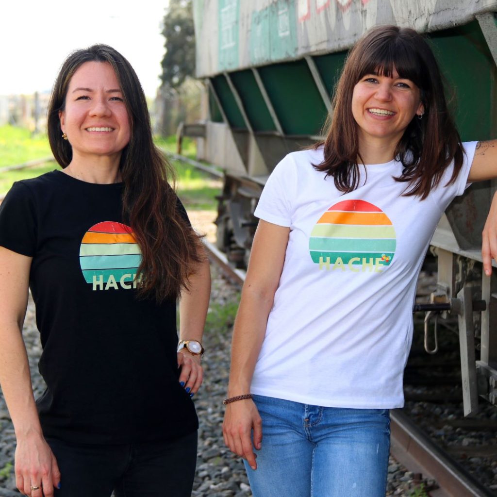 Dos chicas posan con una camiseta con un semicírculo partido en filas de diferentes colores y con la palabra hache en color crema en la fila inferior. Una lleva el modelo negro y la otra el blanco.