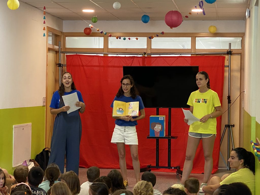 3 de las personas del equipo aparecen en el fondo, de frente, contando el cuento de Bet. En primer plano, los niños y niñas de la ludoteca, de espaldas, atendiendo al cuento.