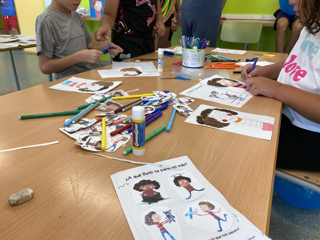 Una mesa llena de rotuladores, tijeras y pegamentos con niños al fondo pintando. En primer plano, un folio donde los niños han pegado diferentes dibujos de Bet.
